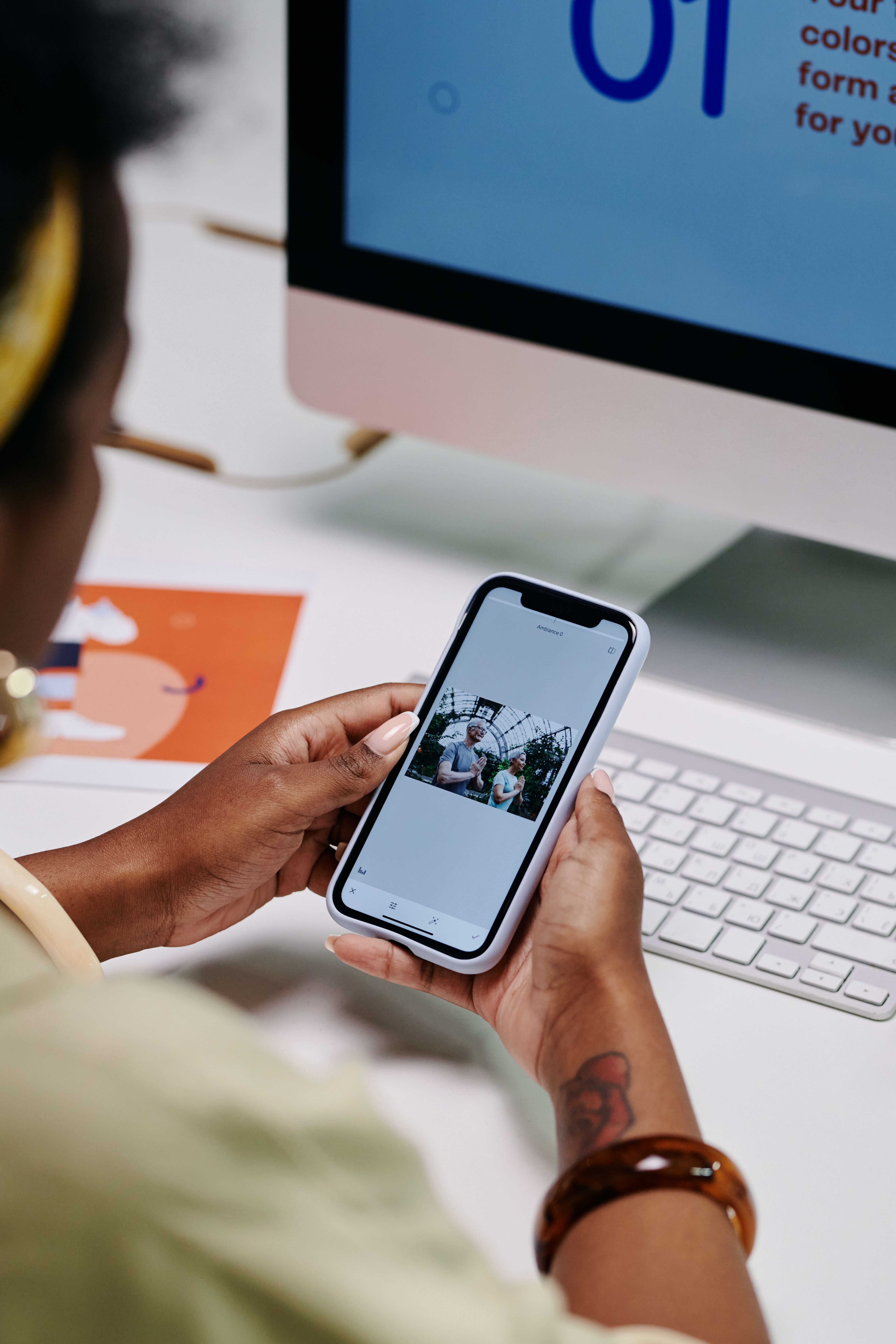 Designer reviewing brand work on a phone beside an iMac showing a color-system layout